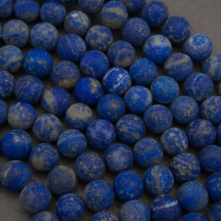 A close-up pile of round blue lapis lazuli stone beads with natural white and gray veining and subtle gold flecks, resting on a neutral gray surface.