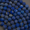 A close-up pile of round blue lapis lazuli stone beads with natural white and gray veining and subtle gold flecks, resting on a neutral gray surface.