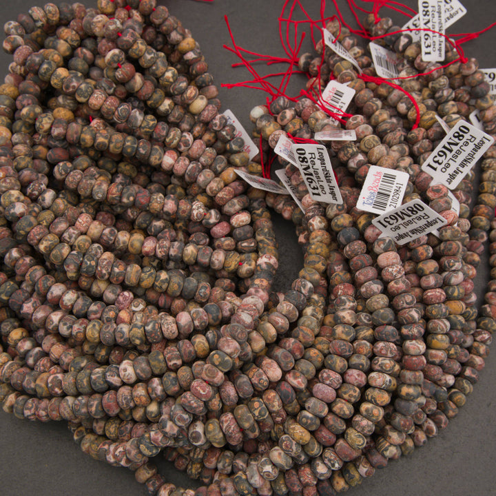 Multiple strands of polished stone beads in earthy multicolor shades, including brown, gray, beige, and muted red, with red thread and white inventory tags attached, arranged on a dark gray surface.