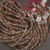 Multiple strands of polished stone beads in earthy multicolor shades, including brown, gray, beige, and muted red, with red thread and white inventory tags attached, arranged on a dark gray surface.