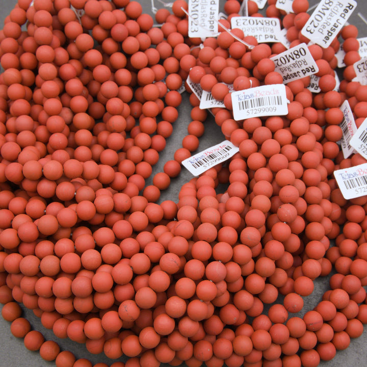 A pile of small matte terracotta red round beads with white product tags attached, photographed on a gray surface.