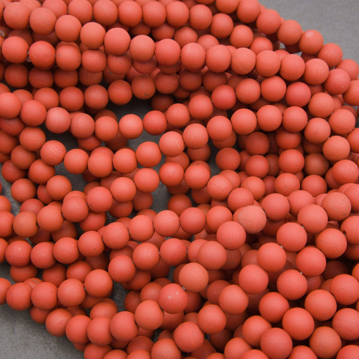 A pile of matte terracotta orange-red round beads or pom-poms filling most of the frame, with a small gray surface visible in the lower left corner.