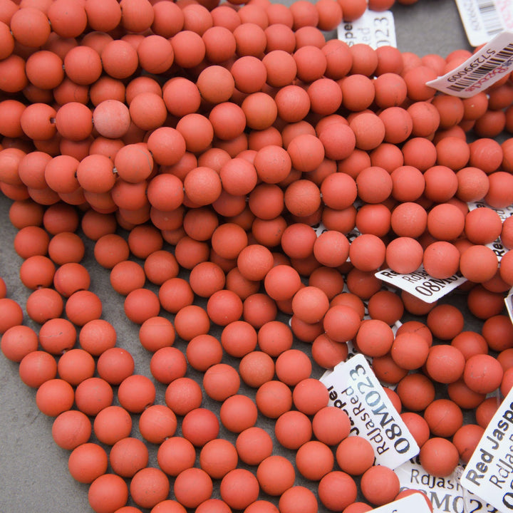A close-up pile of small round coral-red beads with some white inventory tags visible on a gray surface.