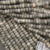 A close-up pile of small cylindrical beads with a mottled gray, white, and beige stone pattern, with a small product tag visible in the lower right corner.