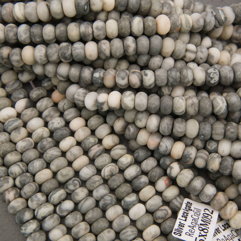A close-up pile of small cylindrical beads with a mottled gray, white, and beige stone pattern, with a small product tag visible in the lower right corner.