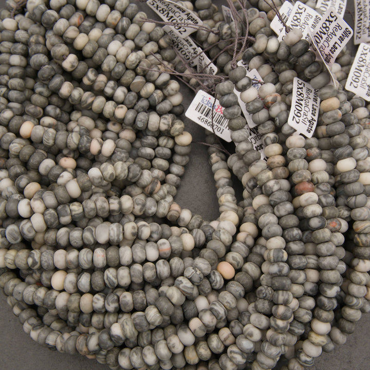 Multiple strands of round stone beads in gray, beige, and white marbled colors, with white inventory tags attached, photographed on a neutral gray surface.