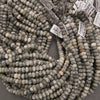 Multiple strands of round stone beads in gray, beige, and white marbled colors, with white inventory tags attached, photographed on a neutral gray surface.