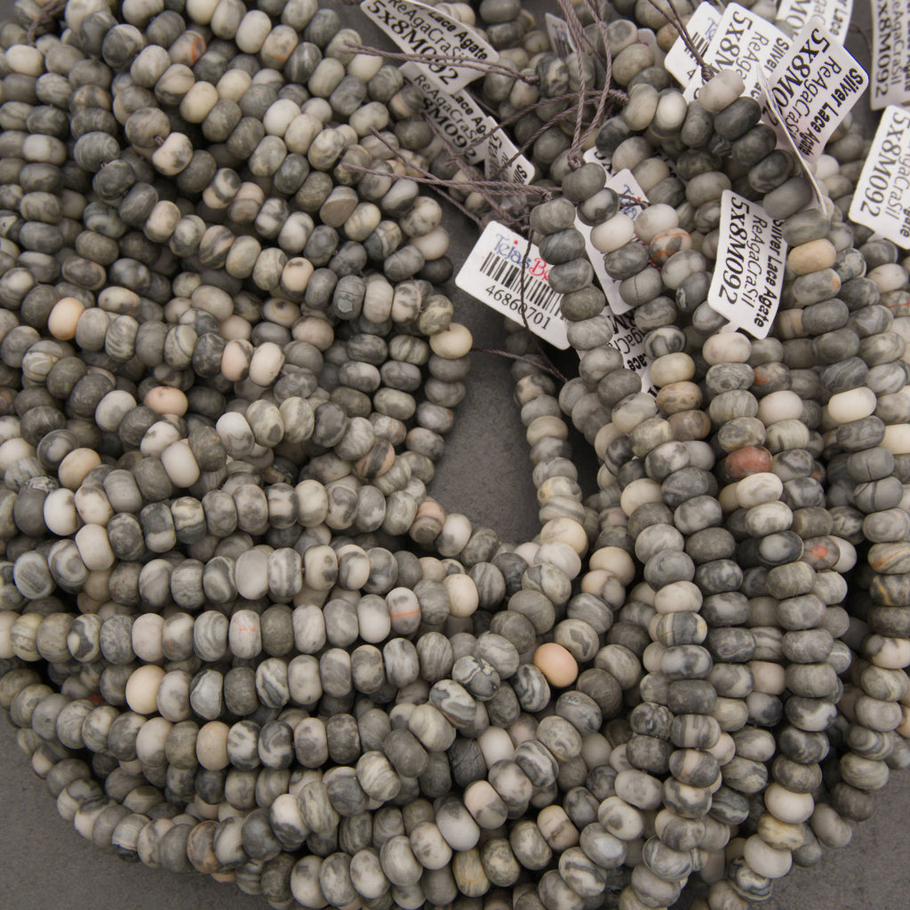 Multiple strands of round stone beads in gray, beige, and white marbled colors, with white inventory tags attached, photographed on a neutral gray surface.