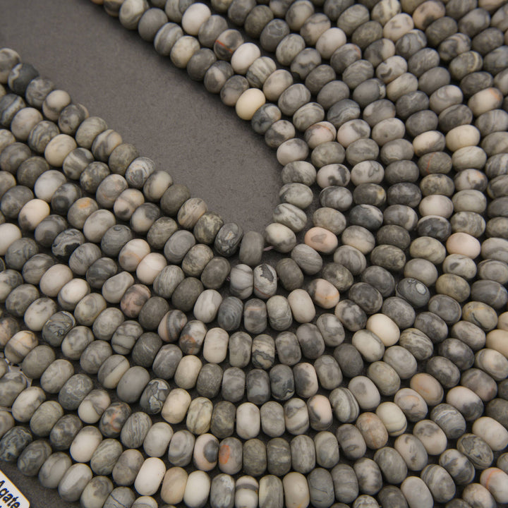 Close-up of multiple strands of small polished stone beads in mixed gray, white, beige, and brown marbled colors arranged on a dark gray surface.