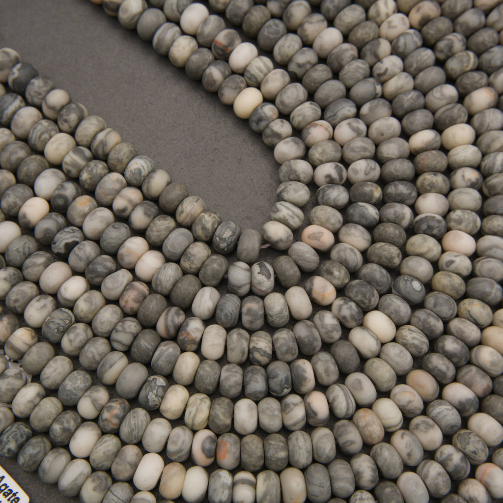 Close-up of multiple strands of small polished stone beads in mixed gray, white, beige, and brown marbled colors arranged on a dark gray surface.