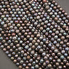 A close-up pile of small round freshwater pearl beads with an iridescent finish, showing shades of dark purple, gray, and green.