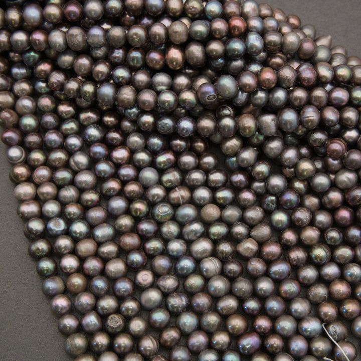 A close-up of a strand of iridescent freshwater pearl beads in a dark gray, bronze, and purple colorway with rainbow reflections, laid out on a dark gray background.