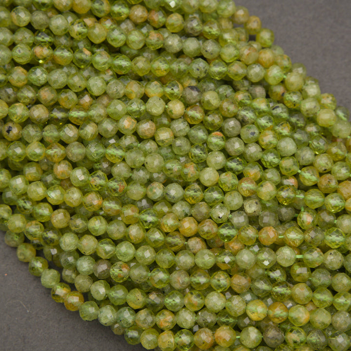 A close-up of many small faceted round beads in a mottled olive green and yellow-green color, piled on a dark gray surface.
