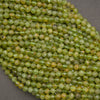 A close-up of many small faceted round beads in a mottled olive green and yellow-green color, piled on a dark gray surface.