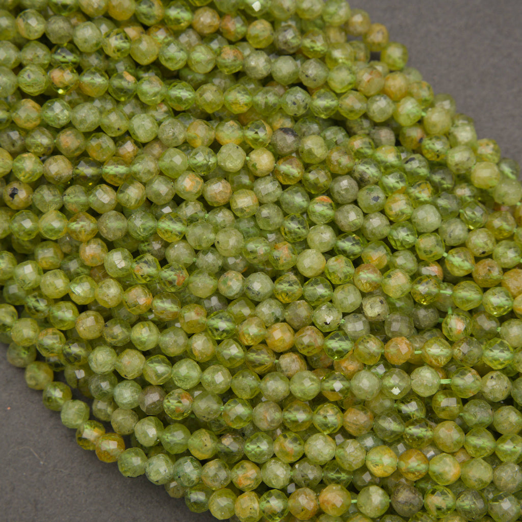 A close-up of many small faceted round beads in a mottled olive green and yellow-green color, piled on a dark gray surface.