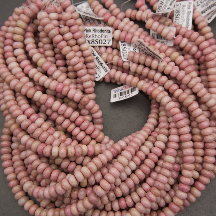 Multiple coils of small round pink rhodonite beads with white product tags, arranged on a dark gray surface.