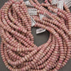 Multiple coils of small round pink rhodonite beads with white product tags, arranged on a dark gray surface.