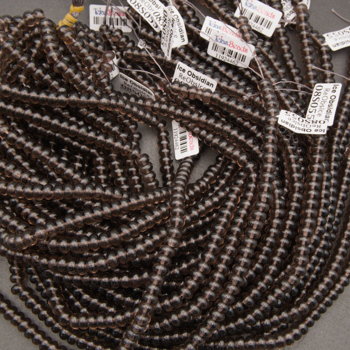 A pile of dark brown translucent gemstone beads with white product tags showing details like 'Ice Obsidian' and 'Cushion Agate'.