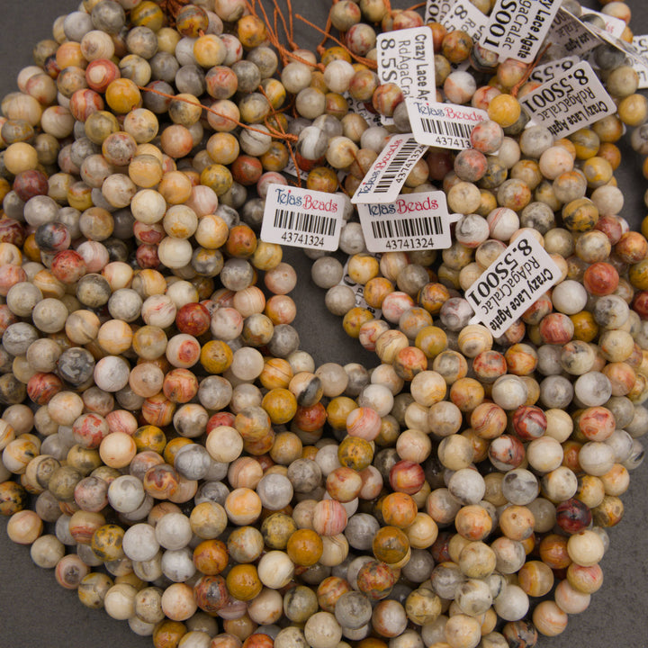 A pile of round gemstone beads in earthy variegated colors, with several white price tags attached. The beads show natural marbling and speckling in shades of beige, brown, orange, gray, and muted red.