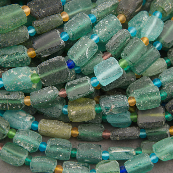 Close-up of a strand of sea glass beads, featuring rectangular and rounded pieces in various shades of green, teal, and gray, separated by small blue and gold spacer beads.