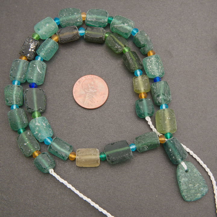 A strand of chunky, rectangular green glass beads with varying shades of emerald and forest green, accented with small blue and gold spacers, displayed on a dark gray background with a US quarter coin for scale.