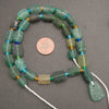 A strand of chunky, rectangular green glass beads with varying shades of emerald and forest green, accented with small blue and gold spacers, displayed on a dark gray background with a US quarter coin for scale.