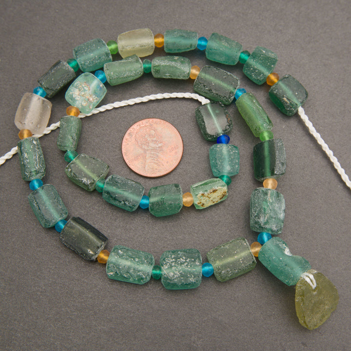 A strand of rectangular green stone beads with small turquoise and gold spacer beads, displayed on a dark gray background with a US quarter coin for scale.
