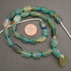 A strand of rectangular green stone beads with small turquoise and gold spacer beads, displayed on a dark gray background with a US quarter coin for scale.