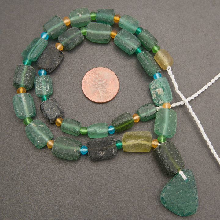 A strand of handmade green glass beads with a large irregular green glass drop piece, displayed on a dark gray background with a copper coin for scale.