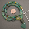 A strand of handmade green glass beads with a large irregular green glass drop piece, displayed on a dark gray background with a copper coin for scale.