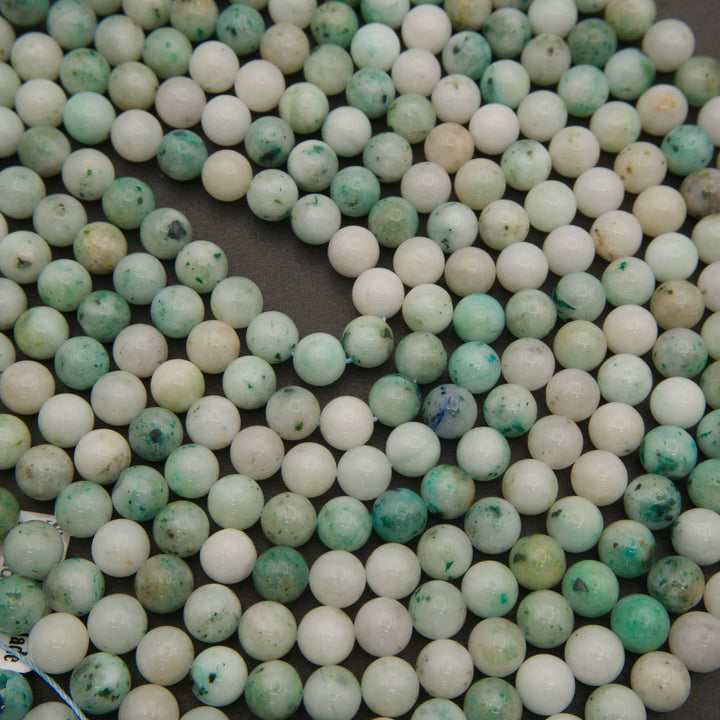Strands of smooth round Sky Mountain Jade beads laid in curving rows on a dark gray background, in white, mint and teal
