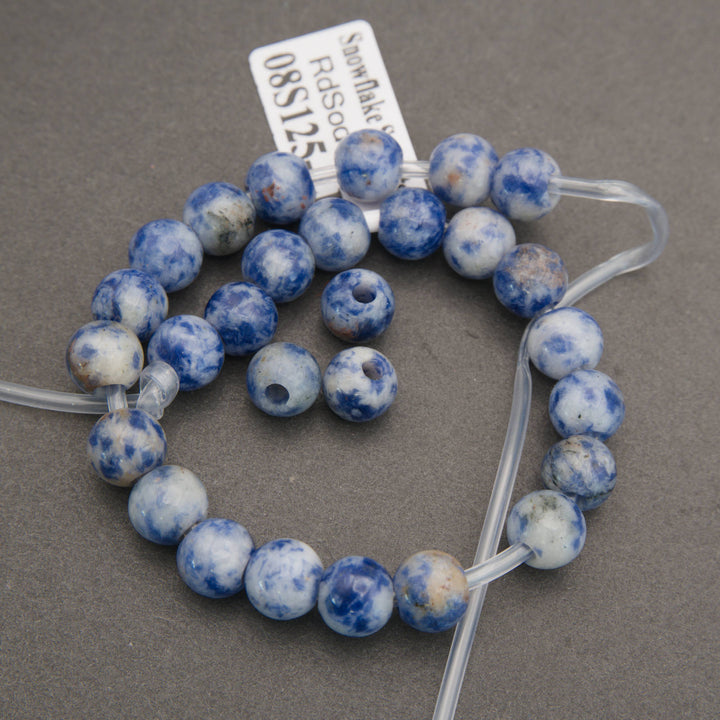 A coiled strand of round blue and white marbled gemstone beads with some natural brownish spots, strung on clear plastic wire with a white tag attached, photographed on a neutral gray background.