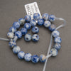 A coiled strand of round blue and white marbled gemstone beads with some natural brownish spots, strung on clear plastic wire with a white tag attached, photographed on a neutral gray background.