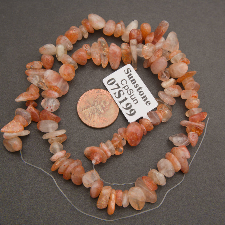 A strand of tumbled orange and peach jasper stone beads on a clear thread, displayed next to a US penny for scale and a white tag labeled 'Sunstone CPSun 07S199' on a dark gray background.