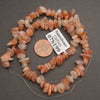 A strand of tumbled orange and peach jasper stone beads on a clear thread, displayed next to a US penny for scale and a white tag labeled 'Sunstone CPSun 07S199' on a dark gray background.
