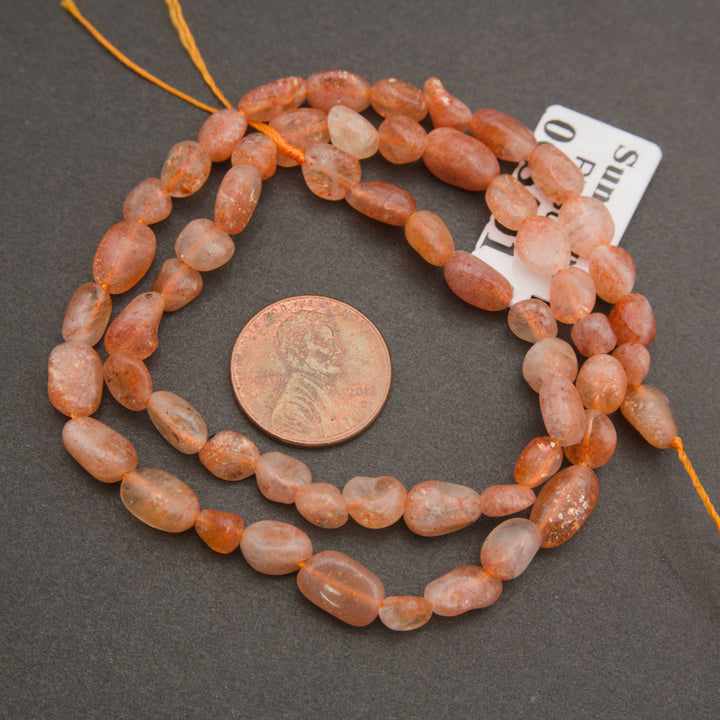 A strand of rough, polished peach-orange and translucent pink gemstone beads, shown next to a US quarter coin for scale and a small white size tag, on a dark gray background.
