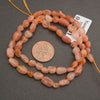 A strand of rough, polished peach-orange and translucent pink gemstone beads, shown next to a US quarter coin for scale and a small white size tag, on a dark gray background.
