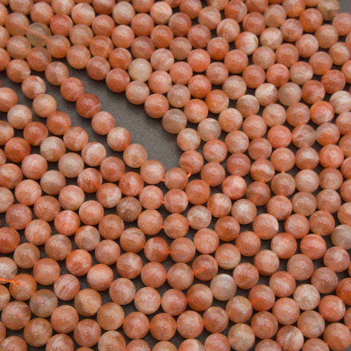 A close-up of many small round beads in a warm peachy orange color with lighter cream marbling, arranged in curved lines over a dark gray surface.