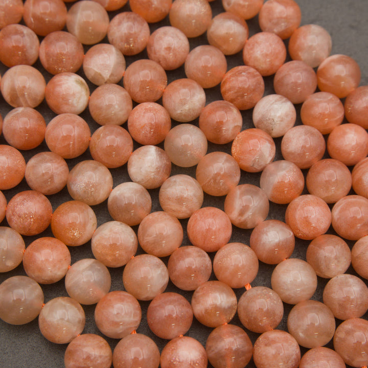 A close-up of many polished round gemstone beads in a peachy orange and translucent pale pink color, arranged tightly together on a dark gray surface.