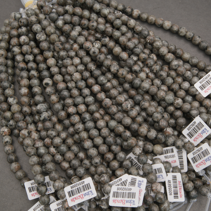 Multiple strands of round stone beads in a speckled gray, brown, and white pattern, with white product tags attached, laid out on a gray surface.