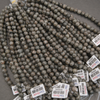 Multiple strands of round stone beads in a speckled gray, brown, and white pattern, with white product tags attached, laid out on a gray surface.