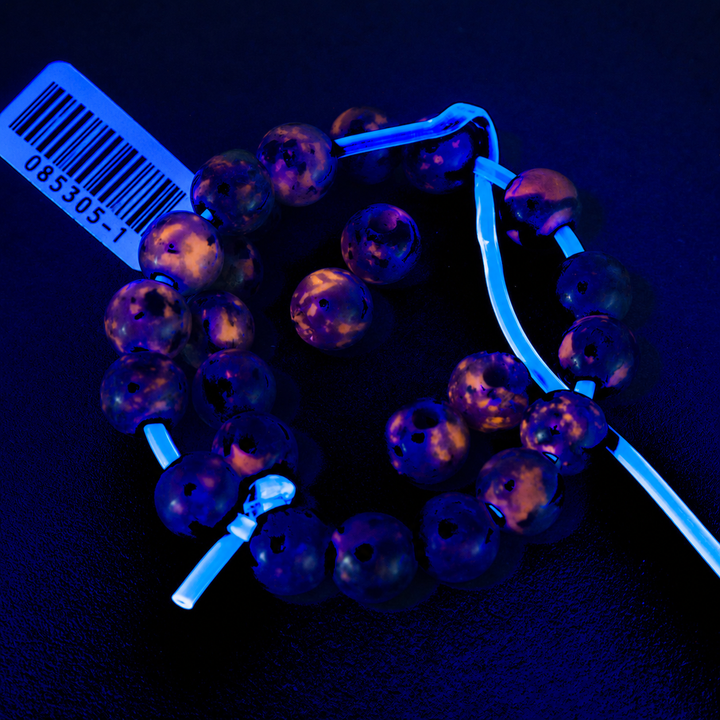 A strand of round translucent beads with a purple and orange marbled pattern, threaded on a light blue cord. A white barcode tag is attached to the strand, and the beads are photographed against a dark, black background under blue light.
