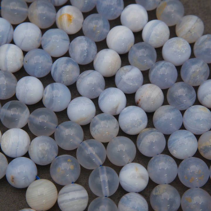A close-up pile of round, polished gemstone beads in pale blue, white, and gray with subtle marbled patterns and a few small warm yellowish inclusions.