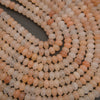 Close-up of multiple strands of small oval beads in soft peach, cream, and light tan tones arranged on a dark gray surface.