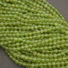 A close-up of many small faceted round beads in a translucent light yellow-green color, arranged in overlapping strands on a neutral gray surface.