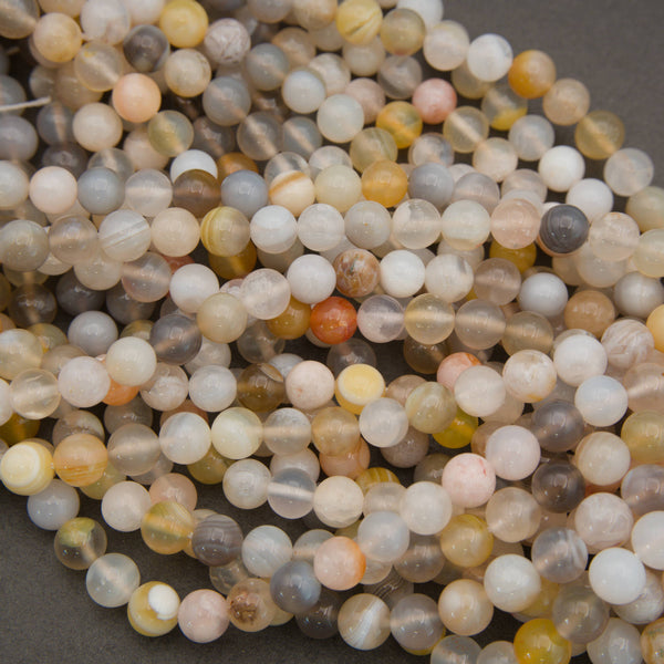 A pile of small round gemstone beads in assorted natural colors and patterns, including white, gray, beige, yellow, orange, and brown, photographed close up on a dark gray background.