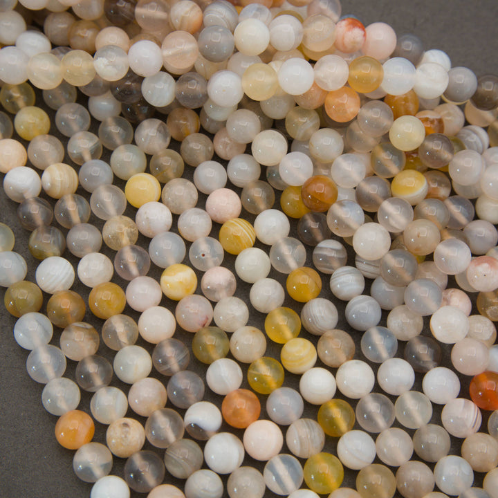 A pile of round, polished agate stone beads in various natural colors and band patterns, including white, gray, yellow, orange, and brown, arranged on a dark gray background.