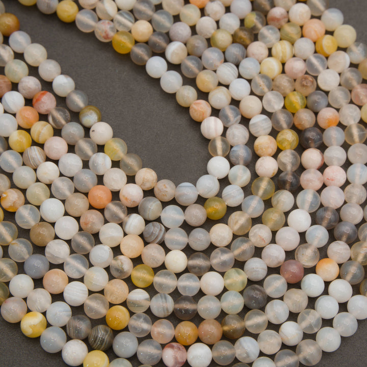 Multiple strands of round polished gemstone beads in a mix of pale gray, cream, white, peach, yellow, and brown tones, arranged in loops on a dark gray background.