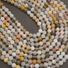 Multiple strands of round polished gemstone beads in a mix of pale gray, cream, white, peach, yellow, and brown tones, arranged in loops on a dark gray background.