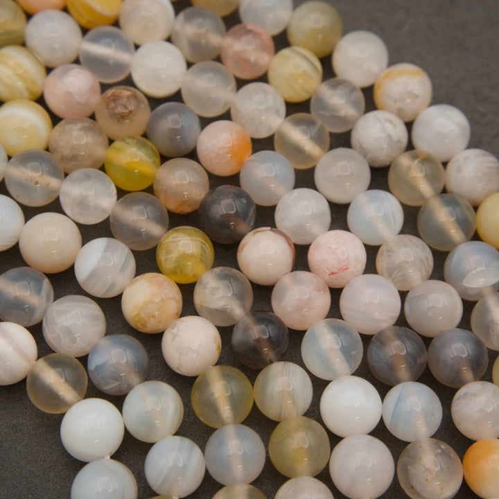 A close-up of many round translucent stone beads in assorted colors and patterns, including milky white, pale pink, soft yellow, smoky gray, and a few darker marbled pieces, arranged on a dark gray surface.